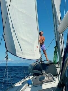 Bernd, der Eigner steht in Badehose an Deck der Indigo unter vollen Segeln bei blauen Himmel.